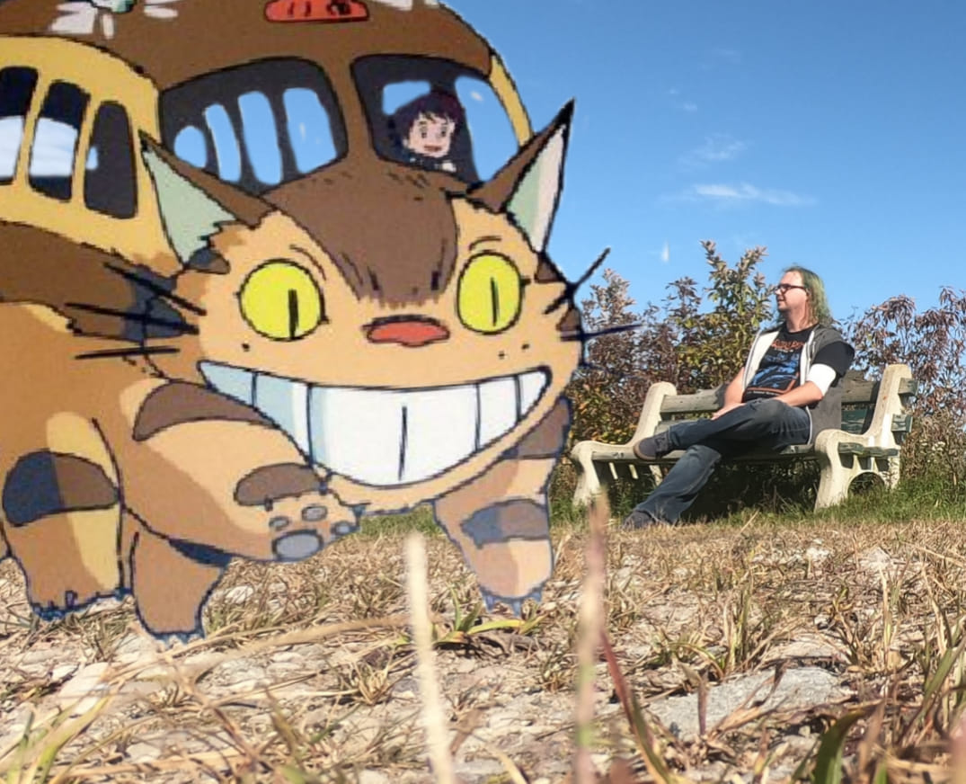 A repeat of the photo of a man sitting on a bench, with a Catbus from My Neighbor Totoro in the foreground